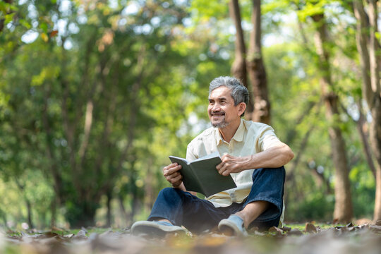 happy asian senior man relaxing in nature park,sitting on grasses and reading a book