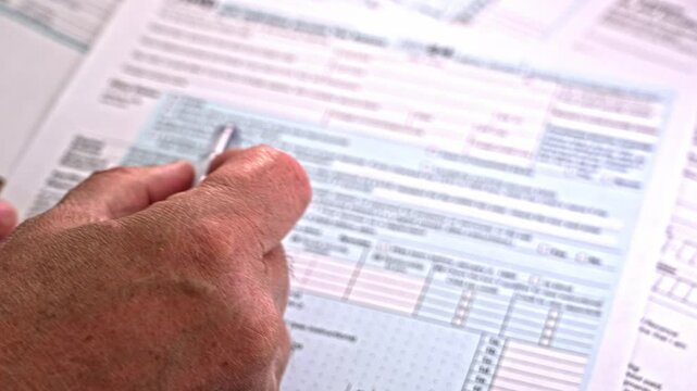A person carefully fills out tax forms at a desk, focusing on financial information. Papers are organized, and a pen is in hand during this detailed preparation process