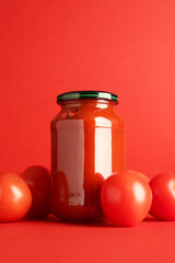 Ketchup in glass jar and fresh tomatoes on red background