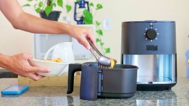 Male hands placing breaded chicken drumsticks into an air fryer. The appliance is on a countertop in a Brazilian kitchen