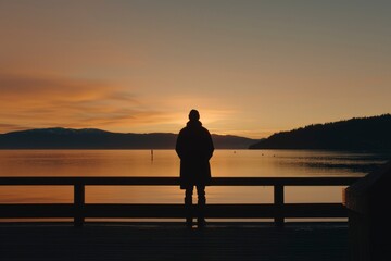 Fototapeta premium Man on dock watches lake at sunrise with mountain backdrop. Travel, reflection