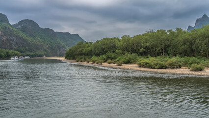 Tourist ships sail along the picturesque river. The riverbed bends. Lush green vegetation on the shores. Mountains against a cloudy sky. China. Li River. Li Jiang. Guilin
