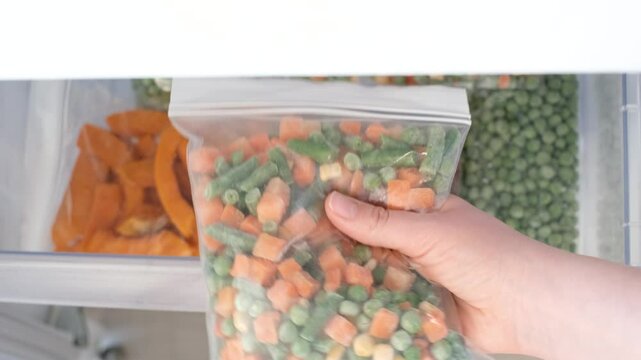 Female hand holding zip bag with frozen chopped vegetables against open freezer background.