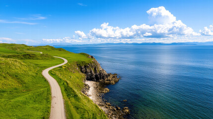 Obraz premium A serene coastal scene featuring a winding path along lush green hills, leading to the tranquil blue sea under a bright sky filled with fluffy clouds.
