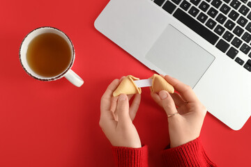 Female hands with broken fortune cookie, cup of tea and modern laptop on red background