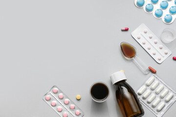 Bottle of cough syrup with measuring cups, spoon and different pills on grey background
