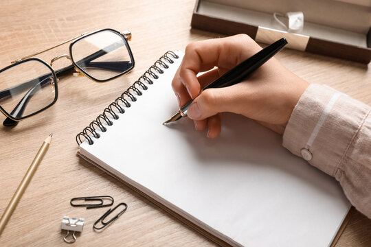 Woman writing something in notebook, eyeglasses, paper clips and pencil on wooden table, closeup