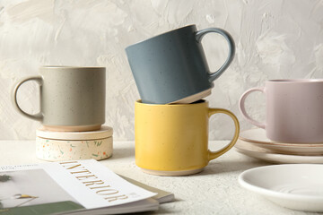 Composition with ceramic mugs and magazines on white table near grey wall
