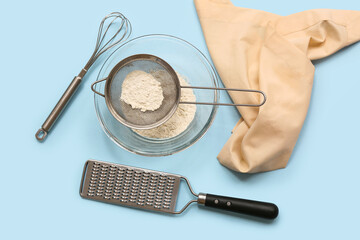 Metal sieve with flour in bowl, grater, whisk and napkin on color background