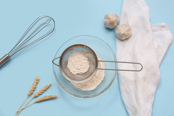 Metal sieve with flour in bowl, whisk, garlic and napkin on color background