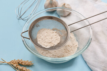 Metal sieve with flour in bowl, whisk, garlic and napkin on color background