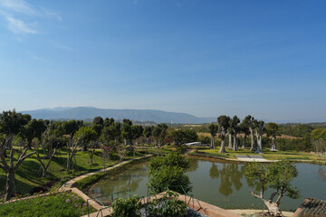 Fototapeta premium A garden with trimmed trees, green lawn, and trimmed bushes in a background of trees with autumn trees on a foot of a blur mountain from afar under a blue sky 