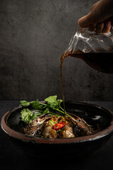 Pouring soy sauce into steamed fish in clay pot on black background