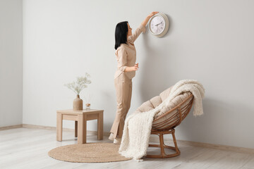 Young woman hanging clock on grey wall in living room. Back view