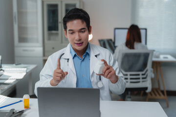 Asian doctor holding a medicine bottle while explaining treatment options to a patient during a video conference in a modern hospital office, utilizing advanced telehealth technology
