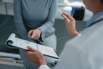 Doctor holding clipboard and pointing at medical chart with pen while explaining diagnosis to patient in hospital room, discussing test results and treatment options