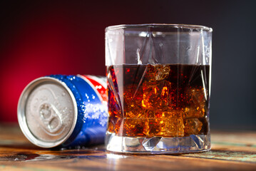 Can of cola and glass of whiskey with ice cubes on red and blue background