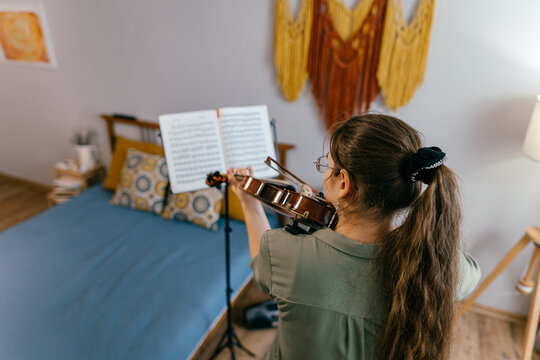 Young woman playing violin at home by sheet music, focused on learning music. Violin student rehearsing classical music in a professional study session