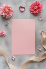 A pink blank paper sheet with a ribbon, flowers, and crystals on a grey concrete table