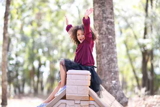 Happy girl with arms raised, sitting atop a playhouse roof outdo