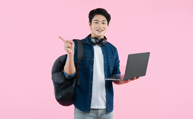 Portrait of Asian male student wearing backpack and headphones , using laptop and  posing on pink...