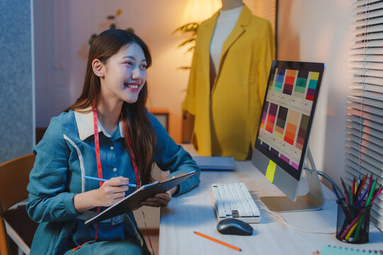 Young fashion designer taking notes on a clipboard while working on new clothing design using a color palette displayed on her computer, with a yellow jacket on a mannequin in the background