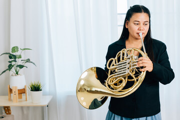 A woman wearing a black jacket holds a French horn in gold, playing it with grace and poise. The...