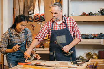 Senior carpenter teaches a woman woodworking techniques, guiding her as she uses a hammer on a wood project. They work together at a well-equipped workbench in a detailed, organized workshop
