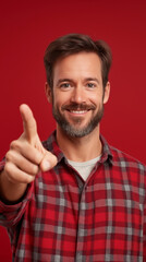 Fototapeta premium Cheerful bearded man in red grey checkered shirt smiling, pointing finger directly at viewer against vivid red backdrop