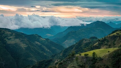 Naklejka premium Scenic mountain landscape with gentle clouds low in the sky during sunset, rich greens in foreground, dramatic blue tones in background mountains.