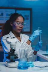 Female scientist wearing lab coat and gloves carefully examining test tube containing blue liquid,...