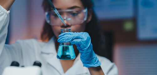 Young female scientist wearing a lab coat and gloves, carefully adding reagent to an erlenmeyer...