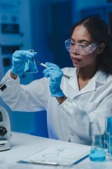 Young female scientist wearing lab coat and gloves, performing experiment with blue liquid in...