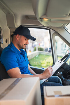 Delivery driver in van recording deliveries with clipboard. Delivery driver concept in a van. Man in blue uniform inside a delivery vehicle, taking notes on a clipboard with packages around him.