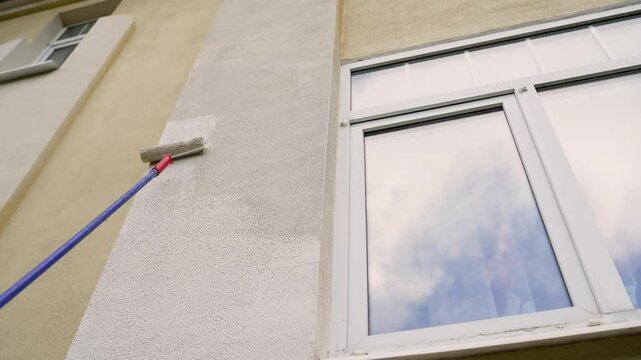 Professional painter smoothing white paint on exterior building facade using roller, carefully maintaining residential property surface with precise painting technique