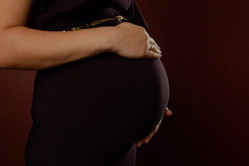 Pregnant woman in black dress hugs her belly with her arms on dark maroon background