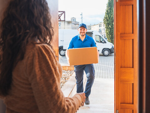 Smiling delivery driver handing package at home doorstep. Delivery driver concept in a van. Man in blue uniform delivering a package to a woman at home, representing a reliable delivery service.