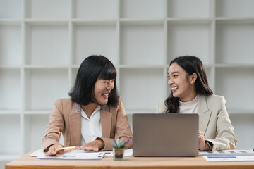 Two cheerful asian businesswomen are enjoying a moment of laughter together while collaborating on a project in a modern office, showcasing teamwork and positive work environment