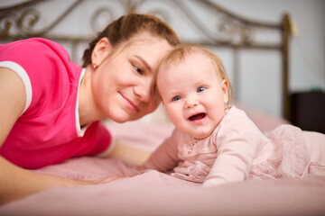 Young woman with brown hair, smiling baby girl indoors. Warm pink tones, soft lighting, metal bedframe in background. Expression of joy and connection