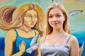 Young blonde female standing confidently before colorful mural. Artwork depicts woman, bird, blue dress. Sunny day, soft shadows enhance details beautifully