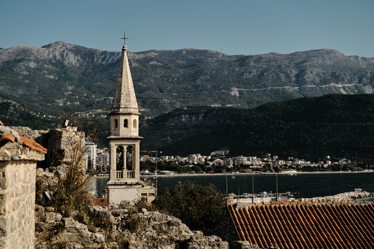Stunning coastal Budva town with a historic bell tower rising above red-tiled rooftops. The calm bay, modern buildings, and majestic mountains in the background.