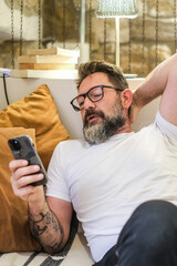Man indoors sitting on his couch in a relaxed manner, looking at his smartphone screen, browsing...