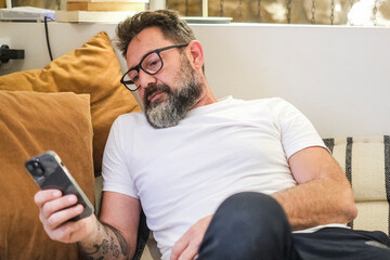 Adult man sitting on a couch at home, looking at his smartphone screen while scrolling through...