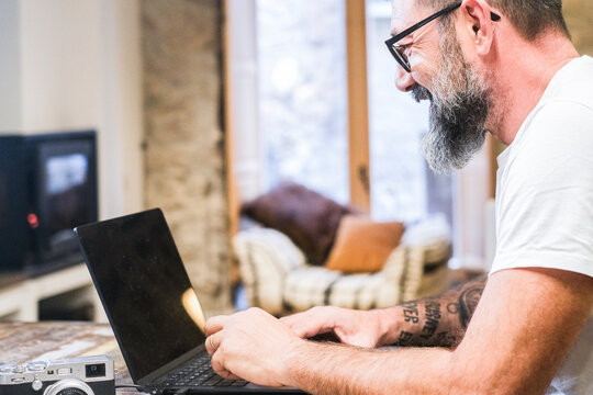 Middle-aged man sitting at home office desk working on laptop with camera on table, living room background with sofa and fireplace, concept of remote work, digital profession, modern workspace