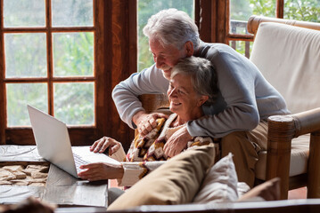 Senior pensioners browsing social media on laptop, woman sitting in chair typing while elderly man stands behind looking at screen, both exploring online tools and staying connected with children