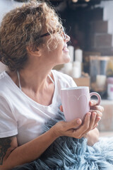 Casual leisure scene indoors with a woman sitting, her curly hair tied up, wearing glasses, holding a pink cup, and keeping a blue furry pillow nearby, highlighting relaxation