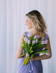 Happy blonde woman holding tulip bouquet purple flowers green leaves white background window light. Joyful girl smiling face enjoys gift. Women's day holiday, march 8, spring concept floral greeting
