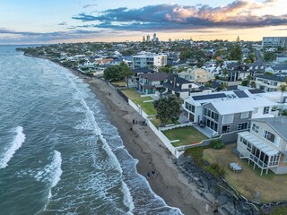 Coastal homes line the beach, with people enjoying the shore. Tranquil scene at sunset. MILFORD, AUCKLAND, NEW ZEALAND