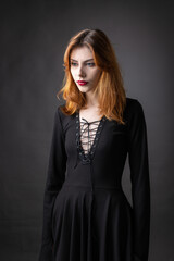 Red-haired gothic girl in a black dress on a gloomy background in the studio.