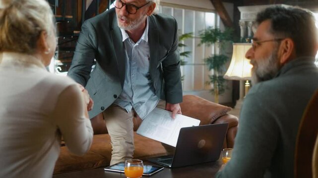 An elderly male insurance agent, sales agent, or real estate agent shakes hands with a retired senior couple after a successful deal at a house meeting. 
